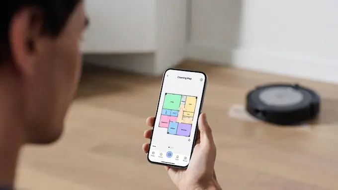 “Hand holding a phone with a Roomba smart cleaning map while the robot vacuums in the background”